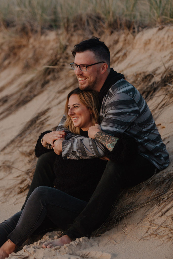 woman sitting in front of man with his arms wrapped around her on a beach