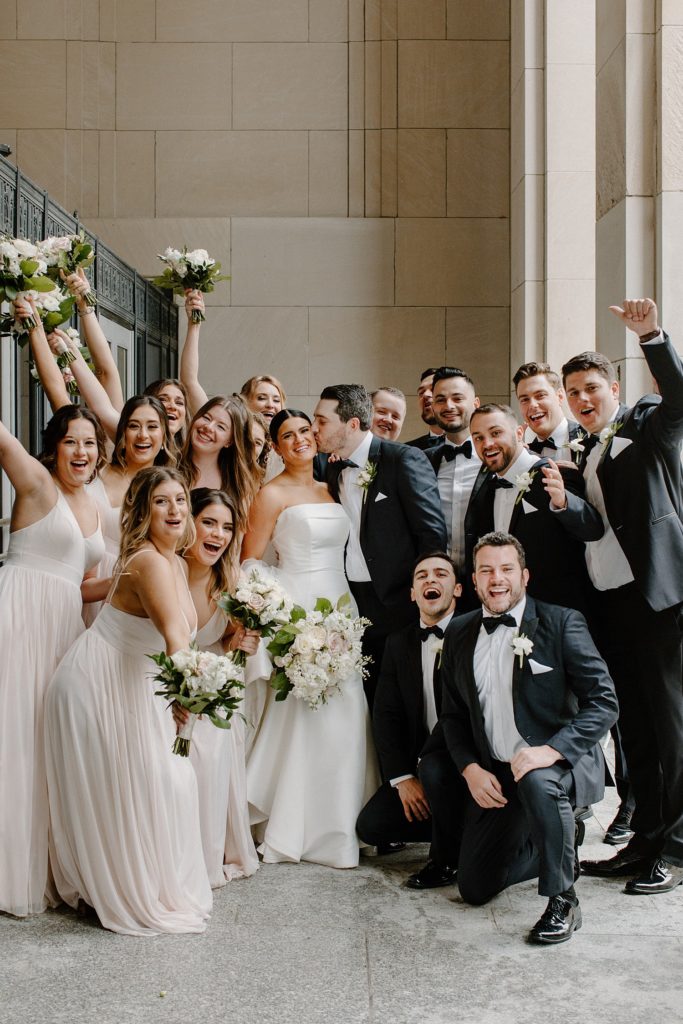 groom kissing bride's cheek while wedding party surrounds them and smiles 