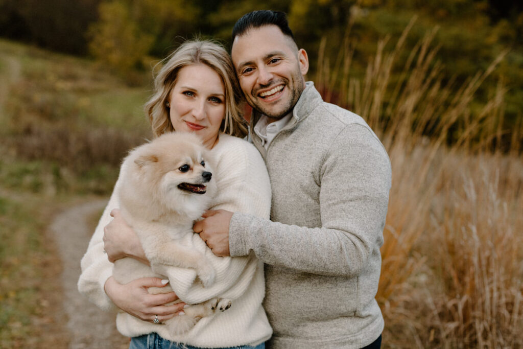 couple standing behind each other smiling as one of them is holding a small dog