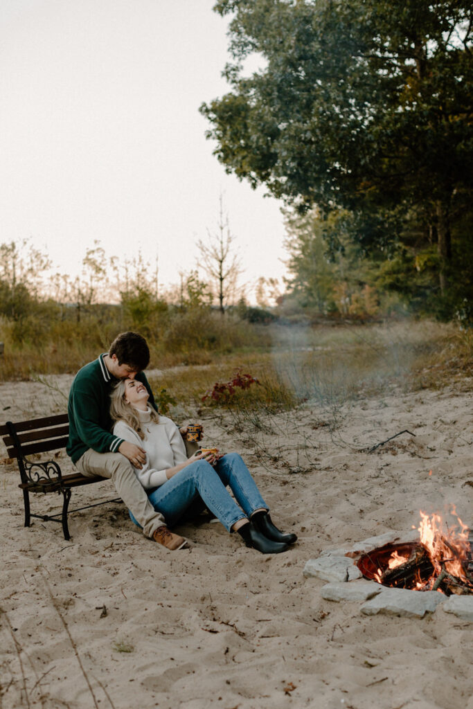 man sitting on a bench by a fire pit with a woman sitting on the sand below between his legs