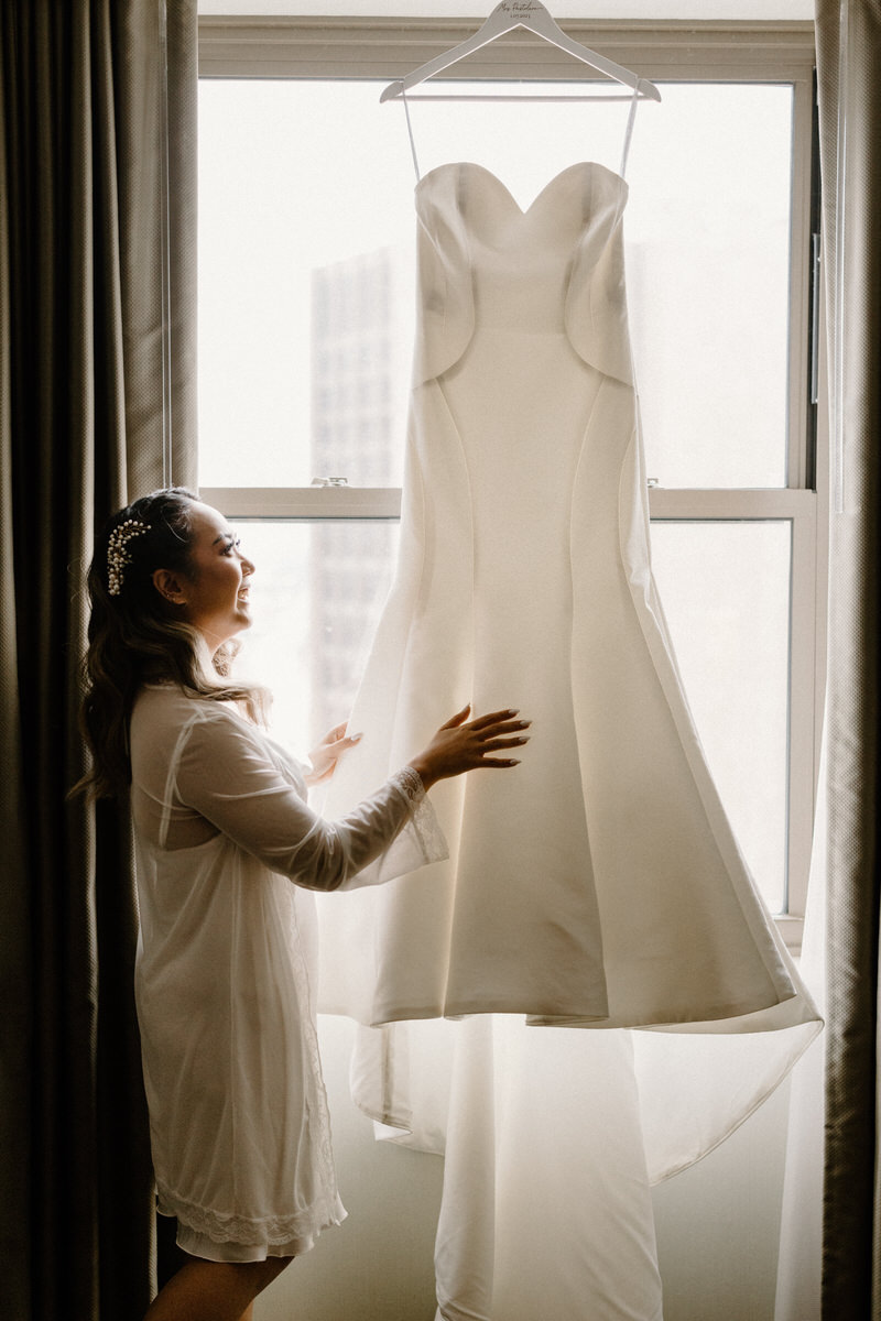 getting ready wedding photo of a wedding dress hanging from a window as the bride touches the bottom of it