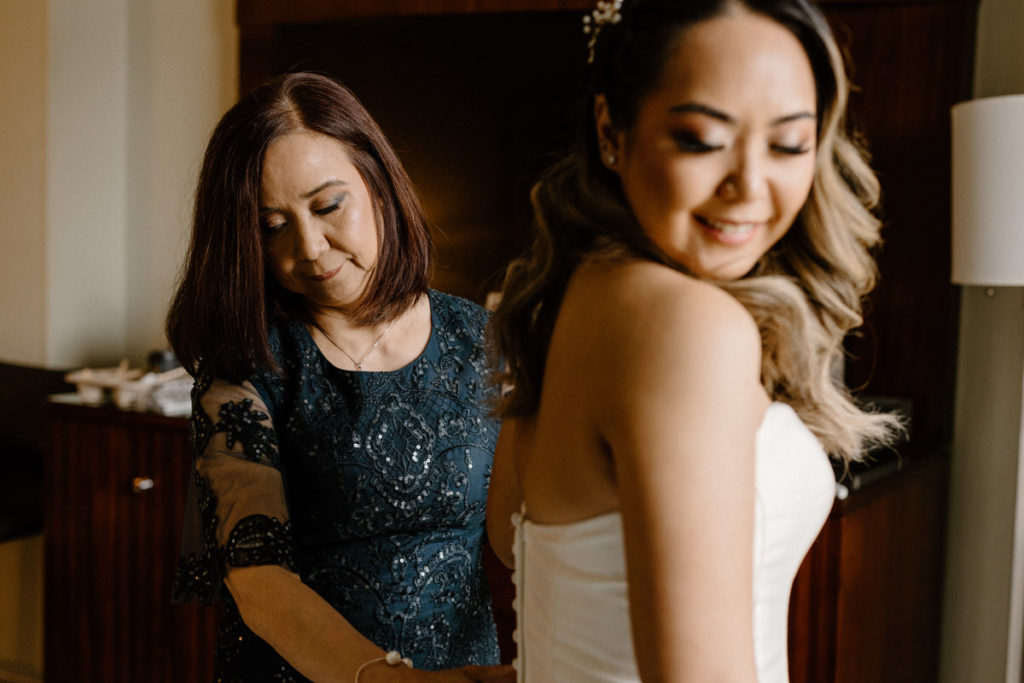 woman helping a bride button up her dress as she looks over her should and smiles