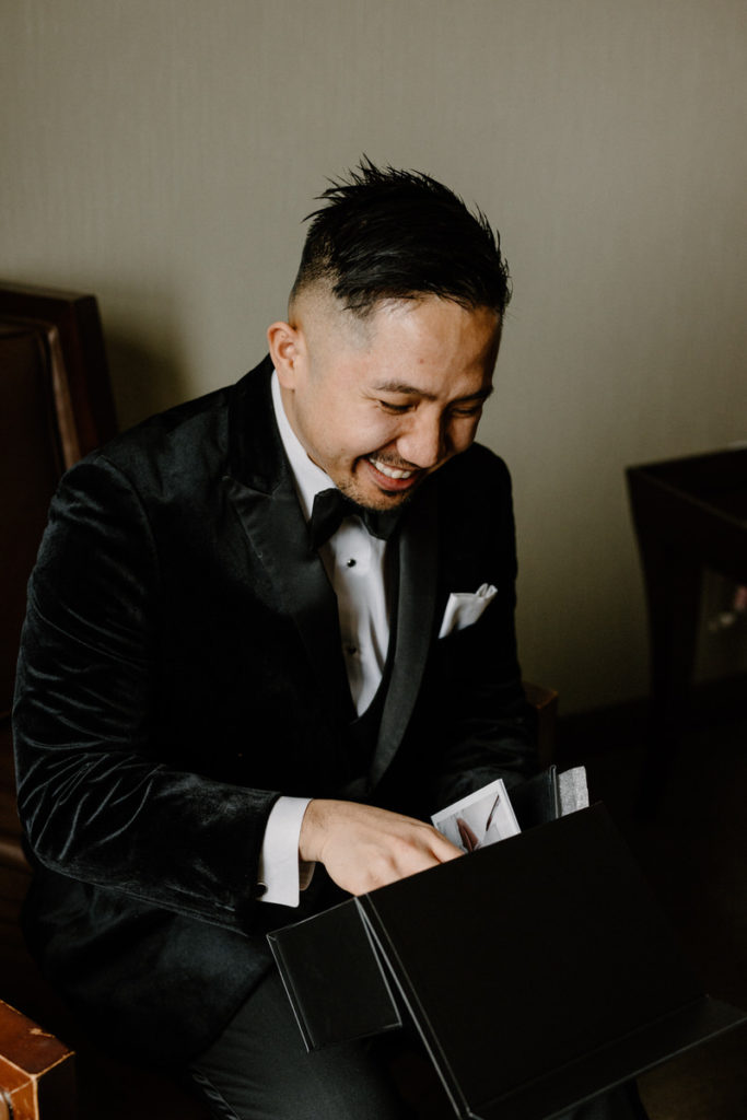 groom sitting down and smiling as he opens a small black box