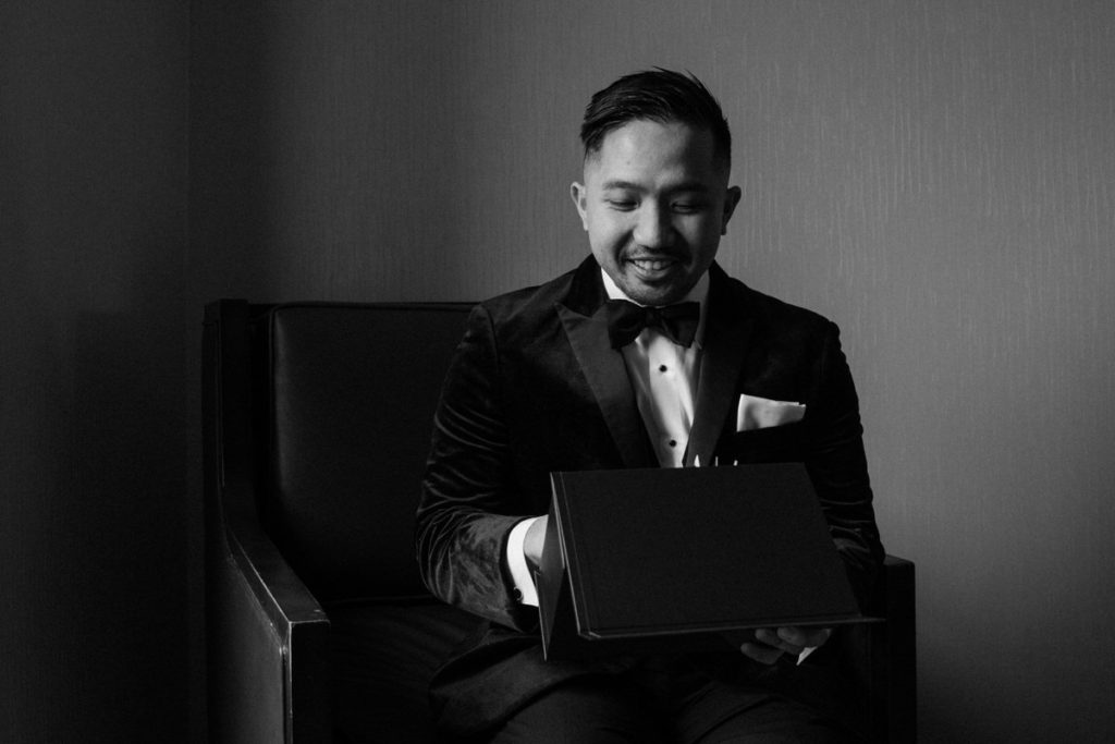 black and white photo of a groom sitting down in a chair opening a black gift box and smiling