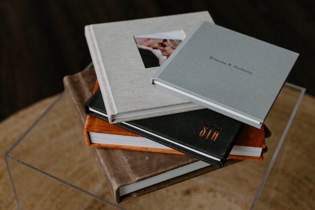 A stack of five wedding photo albums with various colors and material covers including linen, suede and leather, on a clear box.