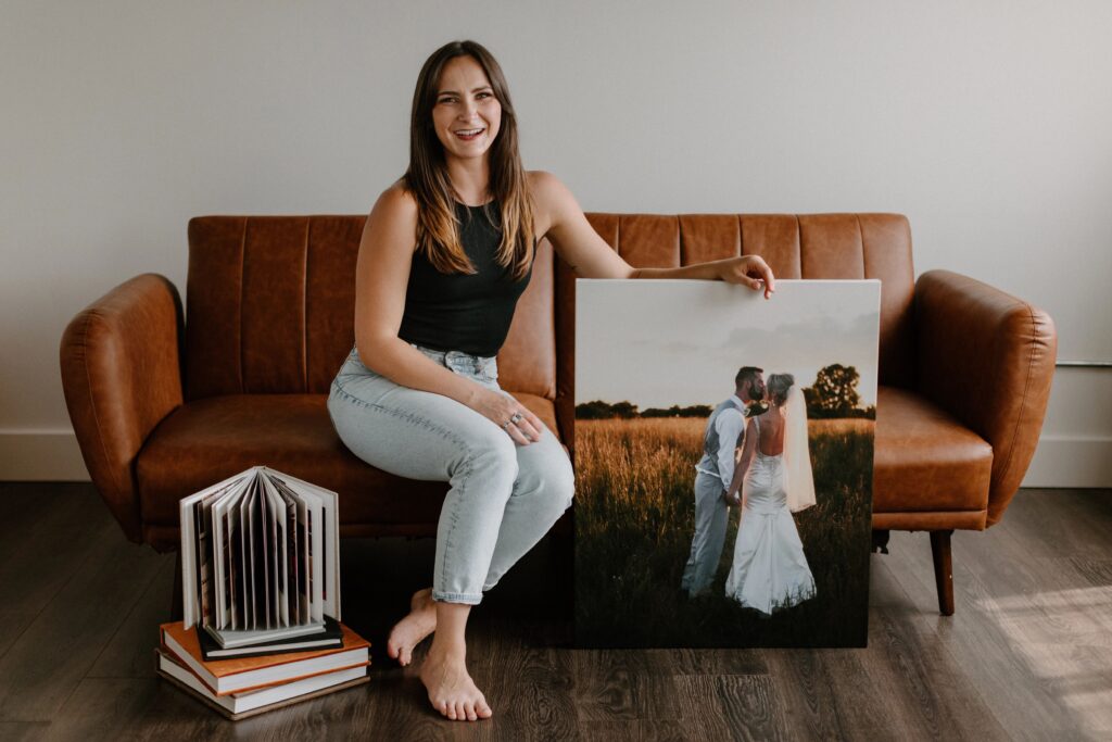 A photographer sitting on a brown leather couch holding up a large canvas photo print of a couple on their wedding day with a stack of wedding albums next to her on the floor.